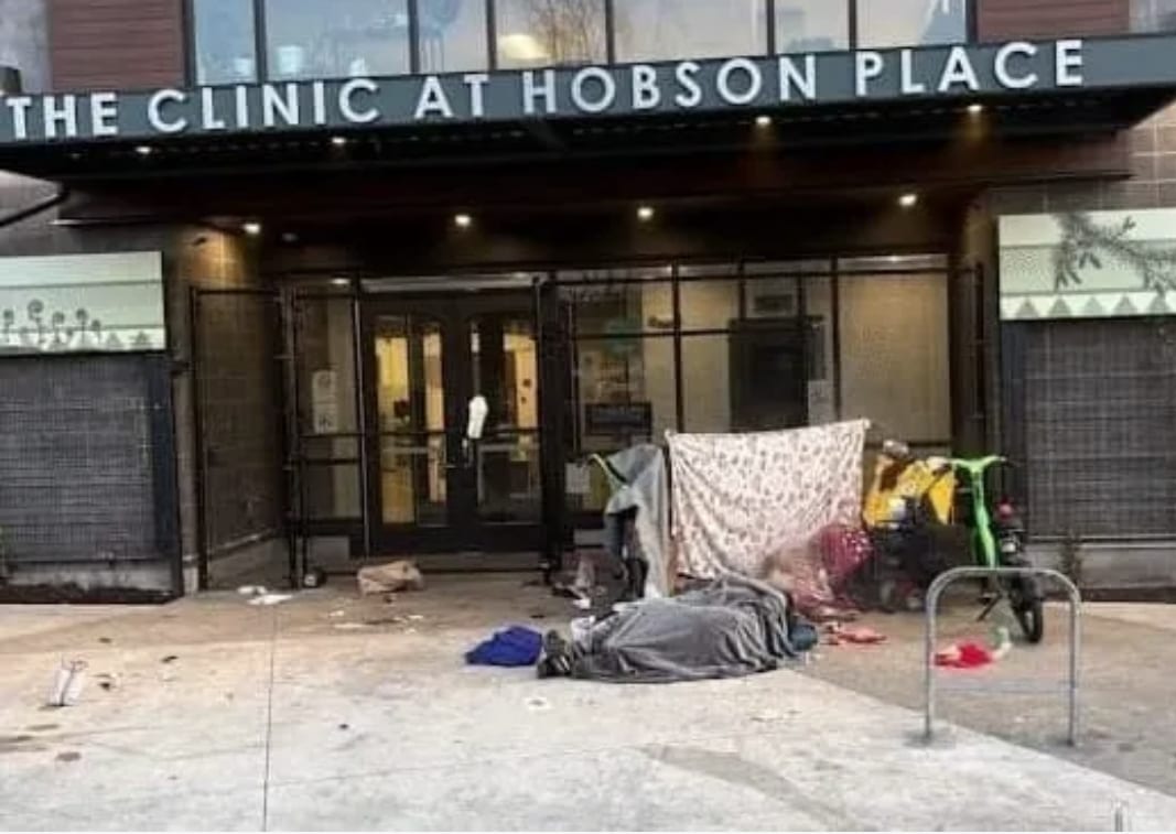 Entrance of The Clinic at Hobson Place, with blankets, belongings, scattered items, and a person lying on the sidewalk