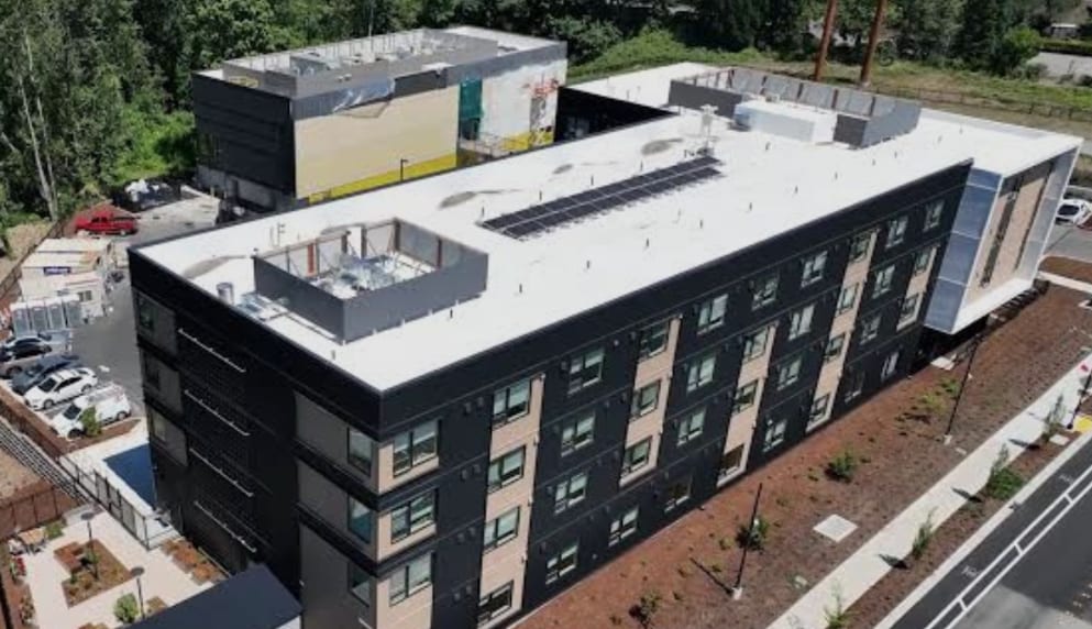 Aerial view of Plymouth Crossing, a dark multi-story building with a white roof and rooftop equipment, beside a parking lot and a neighboring building