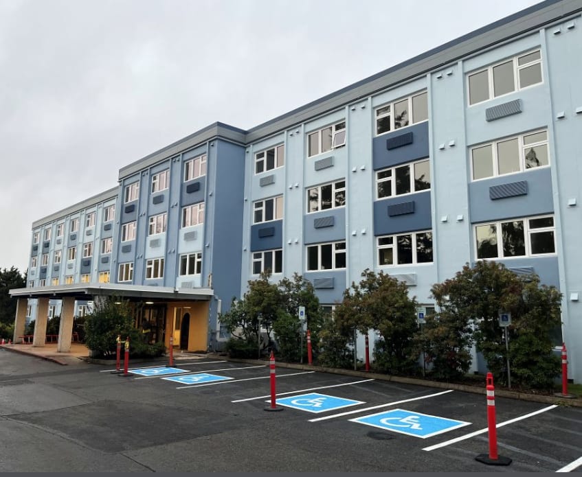 Sheila Stanton Place, a light-blue multi-story building with a covered entry, shrubs, and marked accessible parking in front